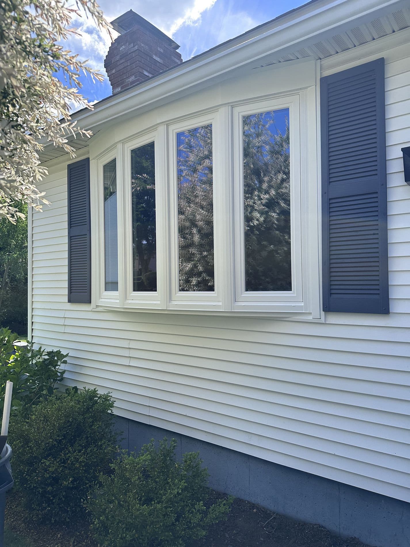 A bow window with white trim and black shutters on a white house above a garden.
