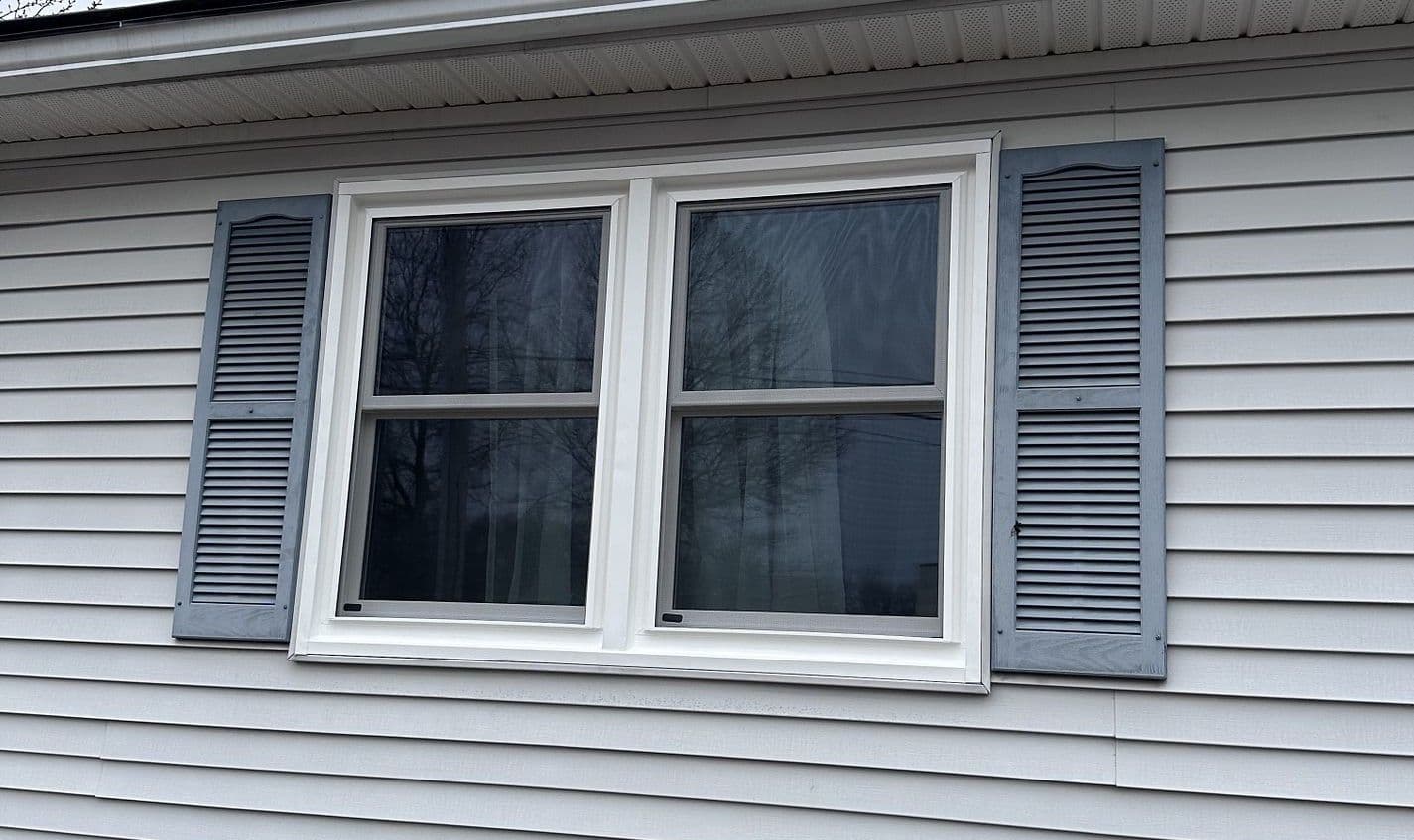 Two double-hung windows with white trim and dark-colored shutters on a light gray house.