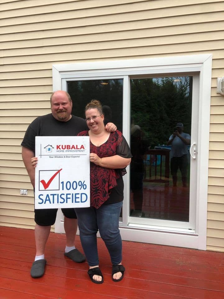 Homeowners hold a sign that reads "100% Satisfied" and stand in front of a newly-installed sliding glass door with white trim on a tan house with a red deck.
