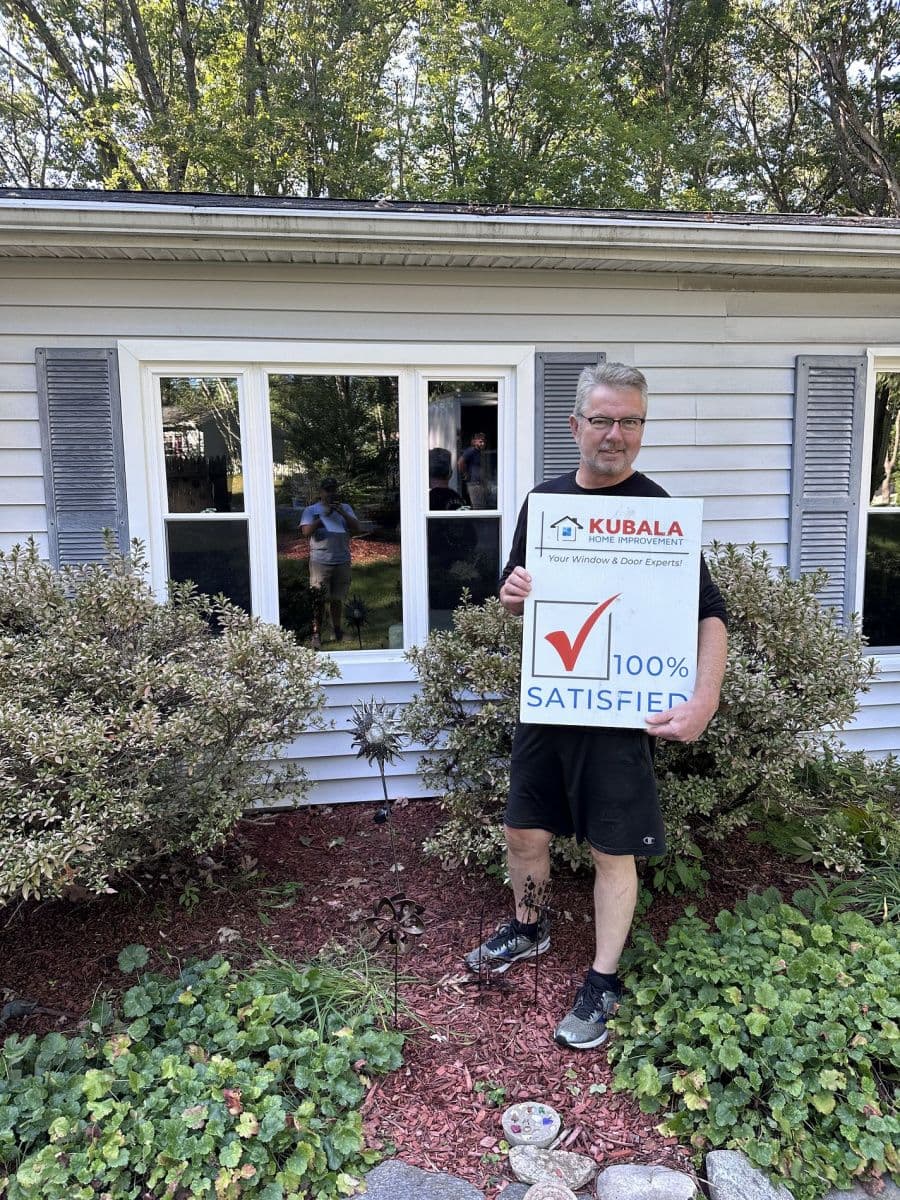 A homeowner holds a sign that reads "100% Satisfied" and stands in front of new replacement windows.