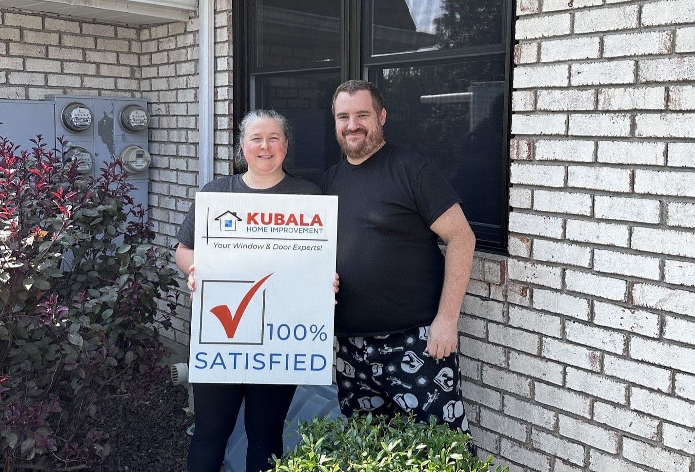Homeowners hold a sign that reads "100% Satisfied" and stand in front of a newly-installed window.